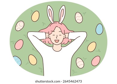 Funny girl with rabbit ears lies on grass among colorful Easter eggs and smiles broadly. Carefree female teenager rejoice at onset of traditional Christian holiday of Easter Sunday