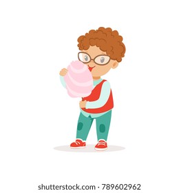 Funny little boy with shiny eyes eating sweet cotton candy. Cartoon kid character with curly red hair wearing glasses, shirt, vest and jeans. Flat vector design