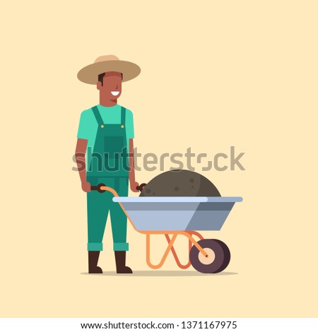 gardener man pushing wheelbarrow full of earth compost african american male farmer working in garden wearing overalls gardening concept full length