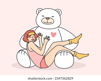 Giant teddy bear near sitting teenage girl in pajamas and sleep mask. Funny schoolgirl teen age looks at screen lying on feet of toy bear rejoicing at cute gift from parents or boyfriend