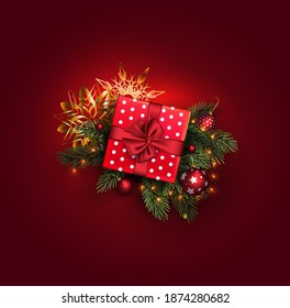 A gift box with a ribbon tied in a bow lies on the branches of a Christmas tree with a garland and decorations.Gold snowflakes on a red background.Concept for a greeting card for the new year