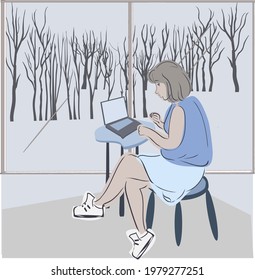 The girl in blue sits at the window, works on a laptop. There is a forest behind the glass.
