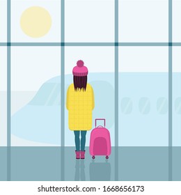 A girl in a bright yellow down jacket and a pink hat with a pink suitcase. A girl in the airport building , looking at the plane through the glass.