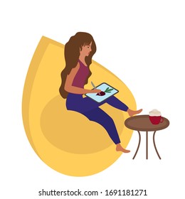 A girl draws an illustration on a tablet. Work from home online, freelance. A comfortable chair and a cup of coffee.