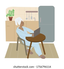 Girl freelancer sits at a laptop in pajamas and with a towel on her head in the kitchen.