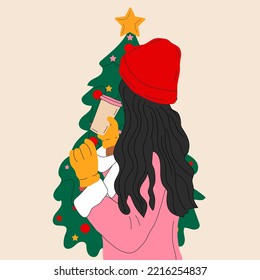A girl in a hat stands in front of a Christmas tree with a cup of coffee