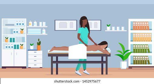 girl having back massage african american masseuse in uniform massaging patient body woman relaxing lying on bed treatments concept modern hospital office interior full length horizontal