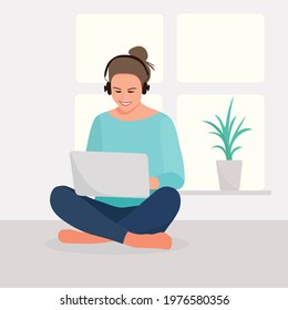 A girl in headphones works at a laptop, a cheerful online dialogue on self-isolation at home. Woman studies on her own near the window in the room, student icon or symbol.