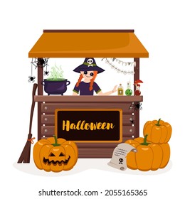 Girl in pointed witch hat at counter of Halloween stall with pumpkins, cauldron, potion and spiders. Joyful child with happy face