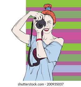 girl with red hair standing and holding a photo camera looks into her narrowed eyes on a striped background