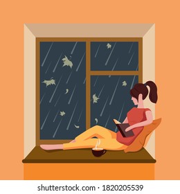 A girl sits by the window, reads a book and drinks tea while it is raining outside the window