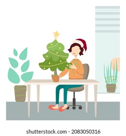 A girl sits in the office at a work table in a red hat and decorates a christmas tree.