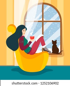 The girl sits at the winter window in a comfortable pillow-chair with a hot cup of drink and reads the story, and a cat sits on the windowsill