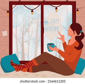 Girl is sitting on the windowsill with a mug of coffee and writes "cozy winter" on the window with her finger. Outside the window is a winter landscape. Vector illustration in flat style