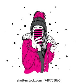 A girl in a stylish down jacket and a beautiful hat with a fluffy bubo. A woman is holding a smartphone and making selfies. Vector illustration. Clothing and accessories, fashion and style.
