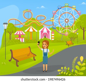 Girl is taking selfie during joyride. Child is photographed on phone camera while walking in park with attractions. Kid with smartphone is making selfie photo against background of amusement park