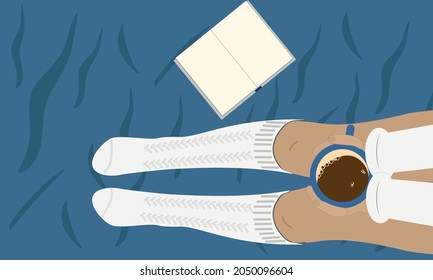 A girl in white socks sits on the bed and drinks coffee and reads a book. Girl on blue crumpled sheet