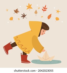 The girl in a yellow raincoat and red boots launches a paper boat. A puddle after a rain. Autumn mood. Leaves fall.
