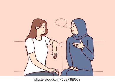 Girls from different ethnicities are talking sitting on couch demonstrating tolerance and lack of cultural prejudice. Woman in muslim clothes is talking to girlfriend of european appearance 