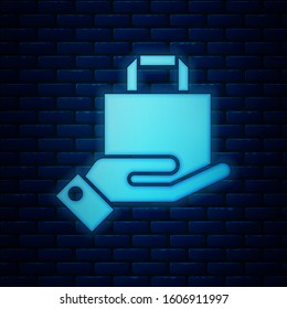 Glowing neon Hand and paper shopping bag icon isolated on brick wall background. Package sign.  Vector Illustration