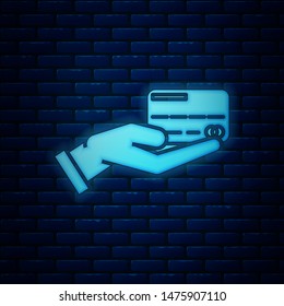 Glowing neon Human hand holding with credit card icon isolated on brick wall background. Online payment. Pay by card. Financial operations.  Vector Illustration