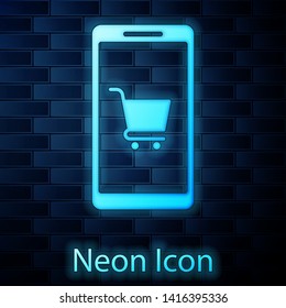 Glowing neon Mobile phone and shopping cart icon isolated on brick wall background. Online buying symbol. Supermarket basket symbol. Vector Illustration