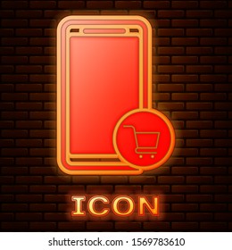Glowing neon Mobile phone and shopping cart icon isolated on brick wall background. Online buying symbol. Supermarket basket symbol.  Vector Illustration