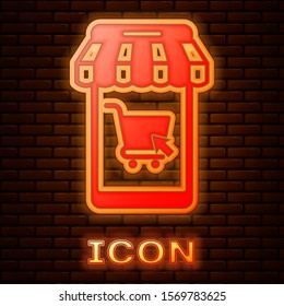 Glowing neon Mobile phone and shopping cart with striped awning icon isolated on brick wall background. Online buying symbol. Supermarket basket symbol.  Vector Illustration
