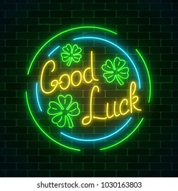Glowing neon sign with geed luck wish and four-leaf clovers in circle frames on dark brick wall background. Hand drawn lettering and three leaves of shamrock. Vector illustration.