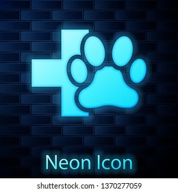 Glowing neon Veterinary clinic symbol icon isolated on brick wall background. Cross hospital sign. A stylized paw print dog or cat. Pet First Aid sign. Vector Illustration