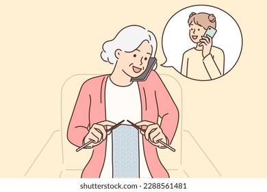 Grandmother makes phone call to granddaughter and knits scarf sitting in chair. Elderly woman with phone is talking to granddaughter, asking how things are at school or inviting to come visit 