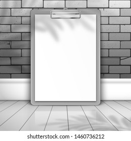 Gray clipboard with white blank paper stands near the brick wall on the boards. The shadow of the leaves of the palm tree is on top. Mockup for design