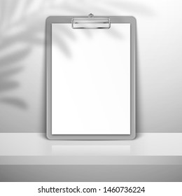 Gray clipboard with white blank paper stands near the wall on the shelf. The shadow of the leaves of the palm tree is on top. Mockup for design