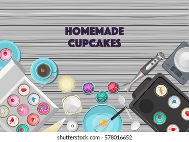 Grey wooden kitchen table with blue ceramic crockery, kitchenware, tools, ingredients and colored homemade cupcakes. Top view.
