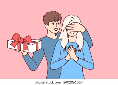 Groom surprises girlfriend standing with gift box and covering girl eyes with hand. Boyfriend in love congratulates happy bride on birthday and Christmas or valentine day on february 14th.