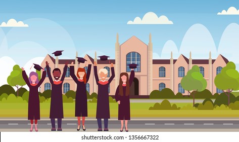 group of mix race students in gown throwing graduation caps in the air standing at campus yard in front of university building exterior education concept flat horizontal full length