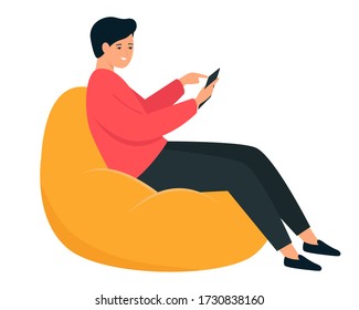 The guy is sitting in a bean bag chair. A young man holds a smartphone in his hand. 