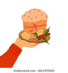 Hand holding a wooden board with traditional Italian panettone bread, decorated with dried citrus slices, holly leaves, red berries, and cinnamon sticks. For Christmas greeting cards, bakery ads