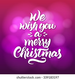 Hand painted brush lettering 'We wish you a merry Christmas' on violet and pink blurred background.