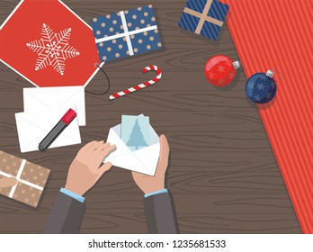 Hands holding Christmas postcard. Christmas presents, balls and gift package on a table. Top view.  Vector Illustration