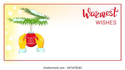 Hands in yellow mittens, gloves holding red Christmas bauble, Christmas ball decoration, hanging on snowy branches of Christmas fir tree, copyspace, lettering Warmest wishes