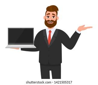 Happy business man in formal wear (suit) holding or showing blank screen of a laptop computer and pointing, presenting something hand palm to copy space. Modern lifestyle, technology device, gadget.