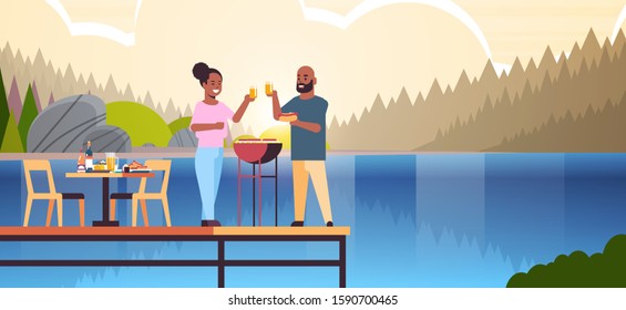 happy couple preparing hot dogs on grill and drinking juice african american man woman in love standing on wooden pier having picnic concept river bank landscape background full length horizontal