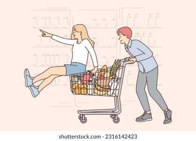 Happy couple walks around supermarket riding food and fruit cart near grocery store shelves. Man rides girlfriend on shopping cart enjoying visiting supermarket with large assortment