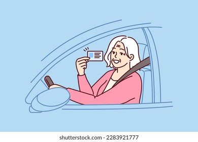 Happy elderly woman demonstrates driver license sitting behind wheel of car and rejoices at opportunity to go on road trip. Positive grey-haired old lady got driver ID after retirement 