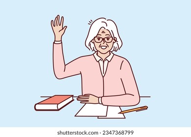 Happy elderly woman student sits at school desk with book and raises hand up wanting to ask question to teacher. Viable positive pensioner student getting education after retirement