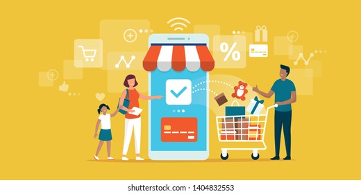 Happy Family Doing Grocery Shopping Online With A Mobile App On Their Smartphone, The Man Is Holding A Shopping Cart With Products And The Woman Is Making The Payment With A Credit Card