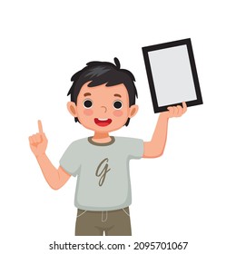 Happy little boy holding up digital tablet with finger pointing up to empty space or copy space for texts, messages and advertising content. Kids and electronic gadget devices concept for children.