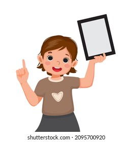 Happy little girl holding up digital tablet with finger pointing up to empty space or copy space for texts, messages and advertising content. Kids and electronic gadget devices concept for children.