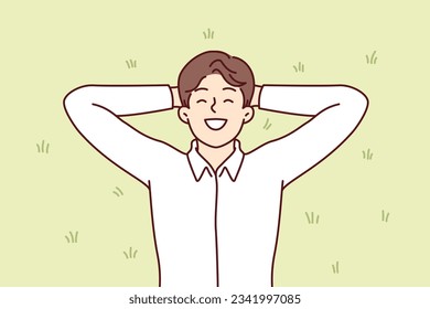 Happy man lies on green grass in park and enjoys warm sunny day and summer weather. Cheerful guy in white shirt with good mood is resting on lawn in park, being energized by sun rays